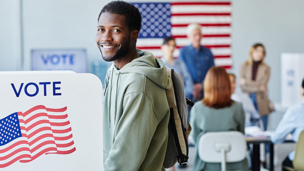 Vote - African American man smiling while voting. American flag and other voters are in the background.